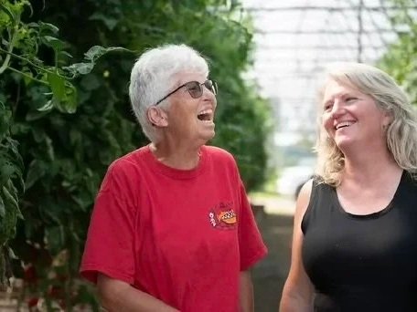 D0D9D852-2EE0-467D-9684-94935D6B64C9 Two women smiling and laughing together in a greenhouse filled with lush green plants, showcasing a friendly and joyful atmosphere. The woman on the left has gray hair and wears glasses, while the woman on the right has long blonde hair.