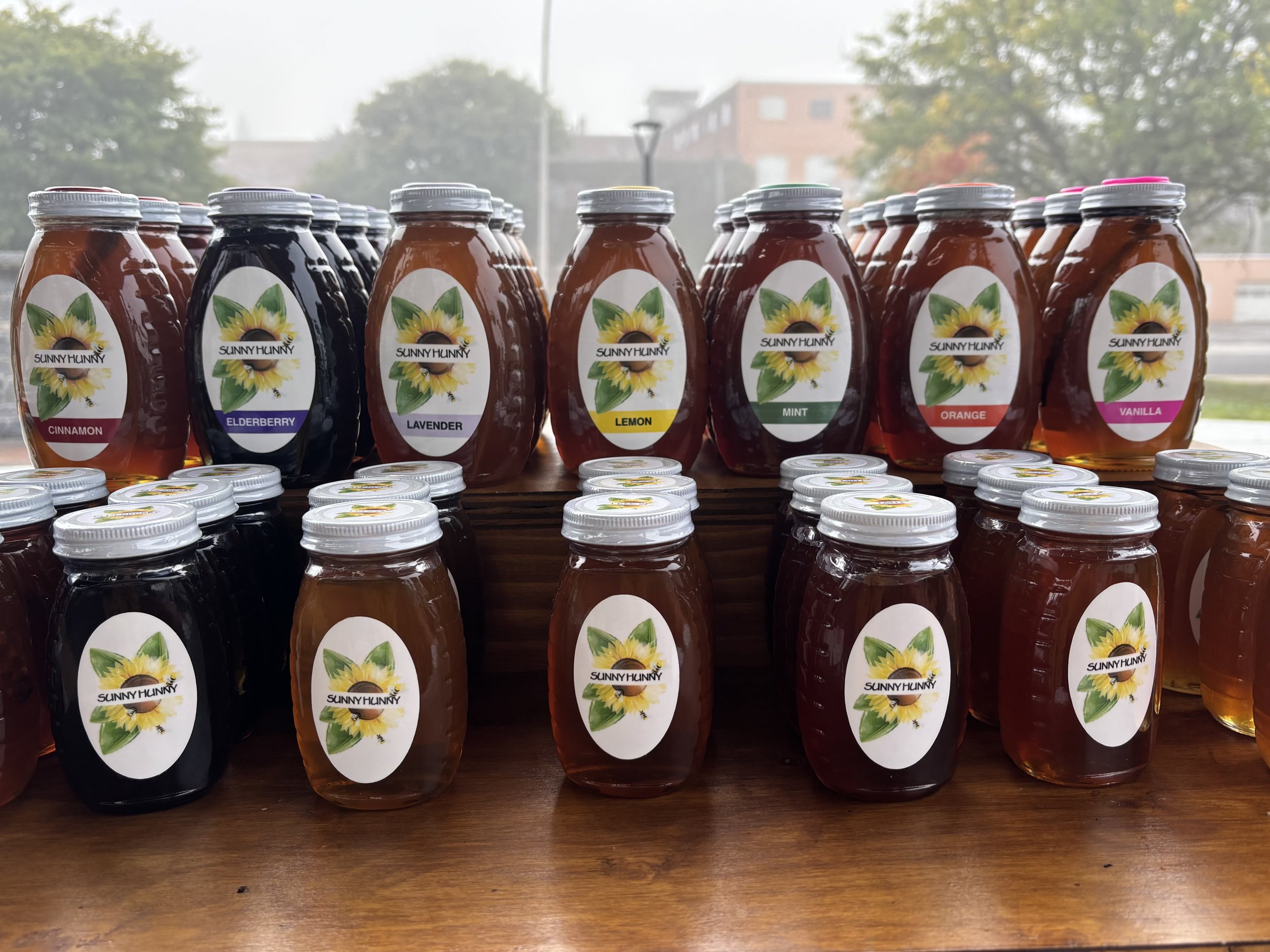 Sunny+Hunny+Farms,+New+York+-+IMG_0909 Assorted jars of Sunny Hunny flavored honey displayed on a wooden table, featuring varieties like cinnamon, elderberry, lavender, lemon, mint, orange, and vanilla, set against a misty backdrop.
